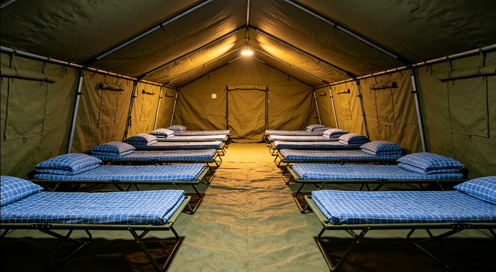 Interior of large military relief tent with rows of camp cots in sky blue checkered bedding, tent poles and canvas walls visible, warm lighting, organized disaster response sleeping arrangement