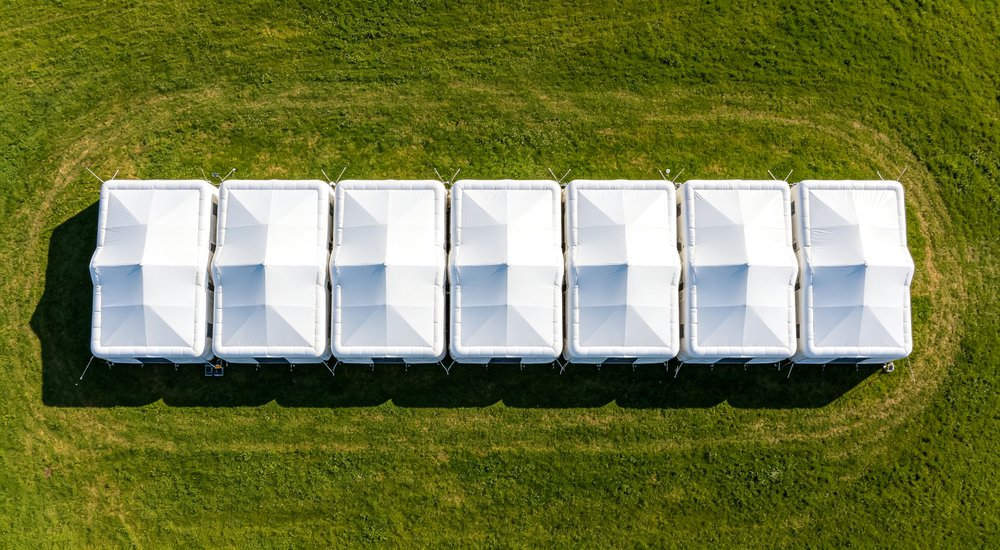 Camp expansion scene with workers adding new module to existing inflatable tent system, green grass, blue sky red and white inflatable emergency shelter for disaster relief operations