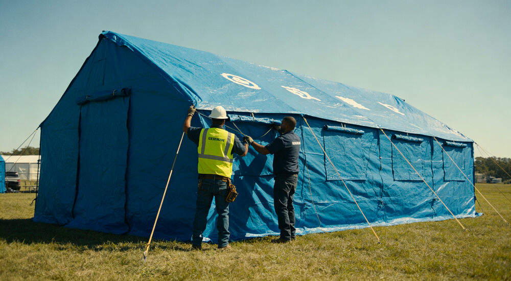 Two workers setting up a large blue relief tent on grass during disaster response
